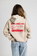 Beiger Oversize-Hoodie mit rotem Aufdruck und Croissant-Grafik, getragen von einer Frau mit offenen Haaren, Hände in den Hosentaschen